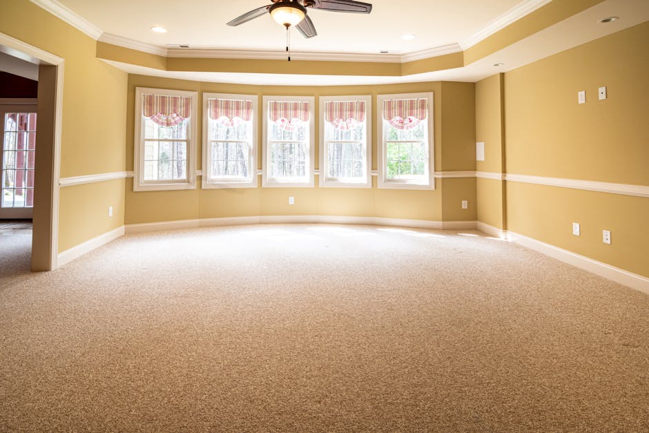 A professional cleaner dressed in full white protective suit, gloves, and mask, performing steam cleaning or deep cleaning of a light beige, textured carpet in a bright, well-lit living room. The room