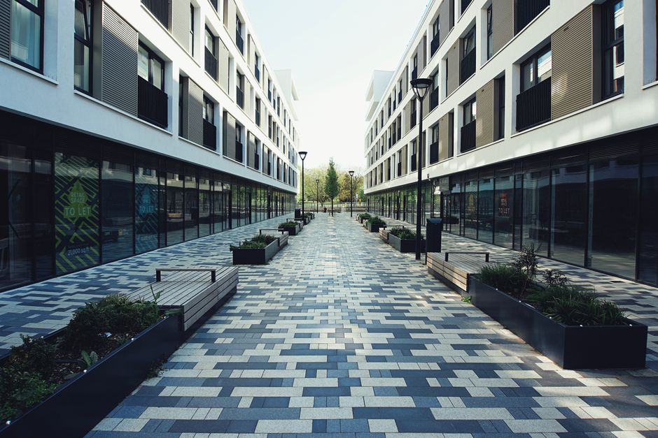Image of a modern outdoor walkway situated between two white multi-storey office buildings with large glass windows. The paved pathway features a patterned surface with rectangular tiles in shades of 