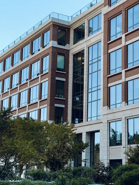 Image of a modern outdoor walkway situated between two white multi-storey office buildings with large glass windows. The paved pathway features a patterned surface with rectangular tiles in shades of 