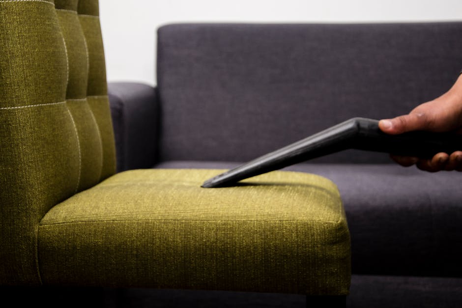 A close-up view of a person using a vacuum cleaner attachment to surface clean a mustard yellow upholstered armchair in a living room, with a dark grey sofa visible in the background. The room has bright, natural lighting, highlighting the fabric texture and cleanliness of the furniture. The scene emphasizes upholstery deep cleaning and surface hygiene carried out by Carpet Cleaners Chiswick, as part of their domestic cleaning services.