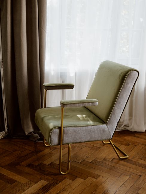 A beige and light green upholstered armchair with gold metal armrests and legs, positioned on a polished wooden parquet floor in front of a large window covered with sheer white curtains and heavier beige drapes. Natural light filters through the curtains, illuminating the clean and well-maintained interior space. The room appears tidy, with no visible dust or clutter, and the overall setting suggests a focus on domestic surface cleaning and maintenance. The scene is associated with professional cleaning services provided by Carpet Cleaners Chiswick, highlighting their expertise in deep cleaning and upholstery sanitisation in a residential environment.